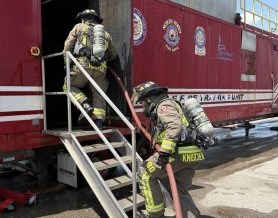 Two firefighters carrying a hose climb stairs to a red fire training facility