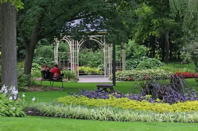 A gazebo in the middle of a park with trees and colourful flowerbeds